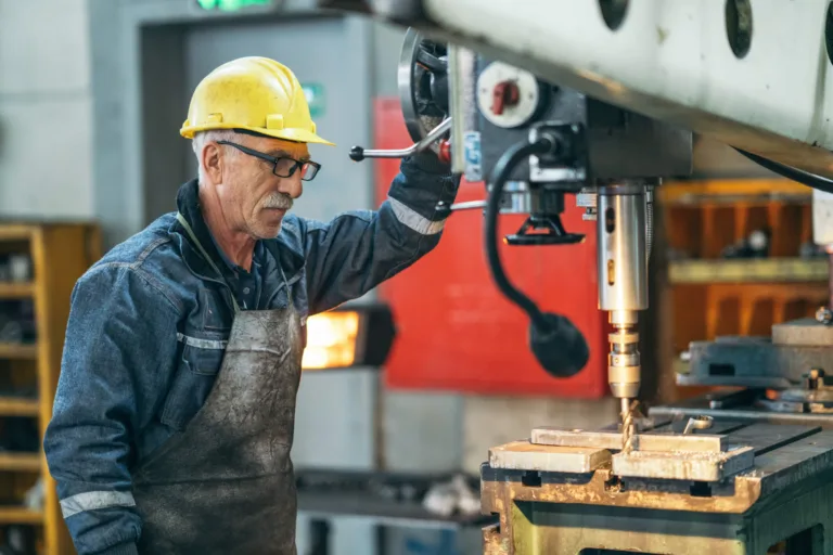 Worker in yellow hard hat operating a drill press in a machine shop