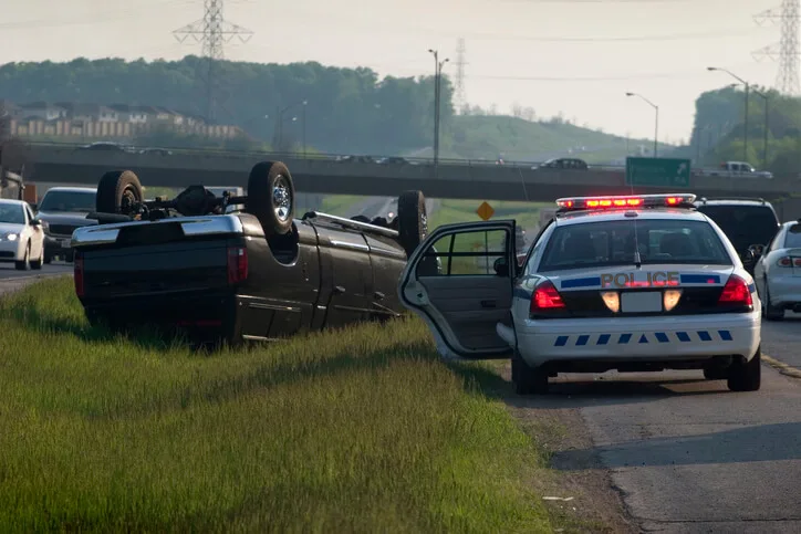 Police at scene of overturned pickup on highway shoulder, lights flashing.